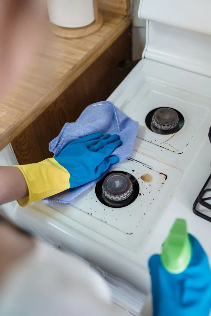A gloved hand wiping a gas hob with a purple cloth next to a green spray bottle