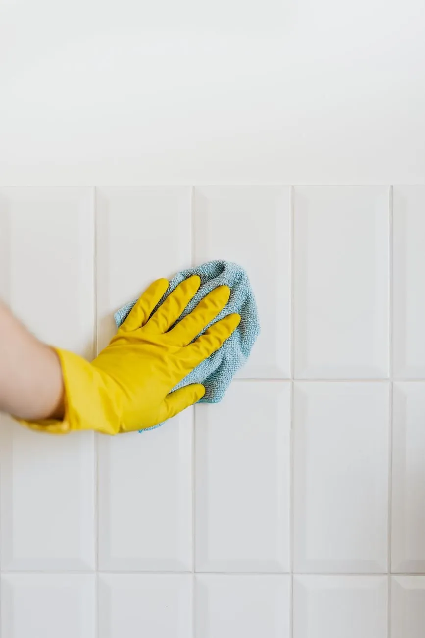 A yellow-gloved hand wiping white wall tiles with a cloth