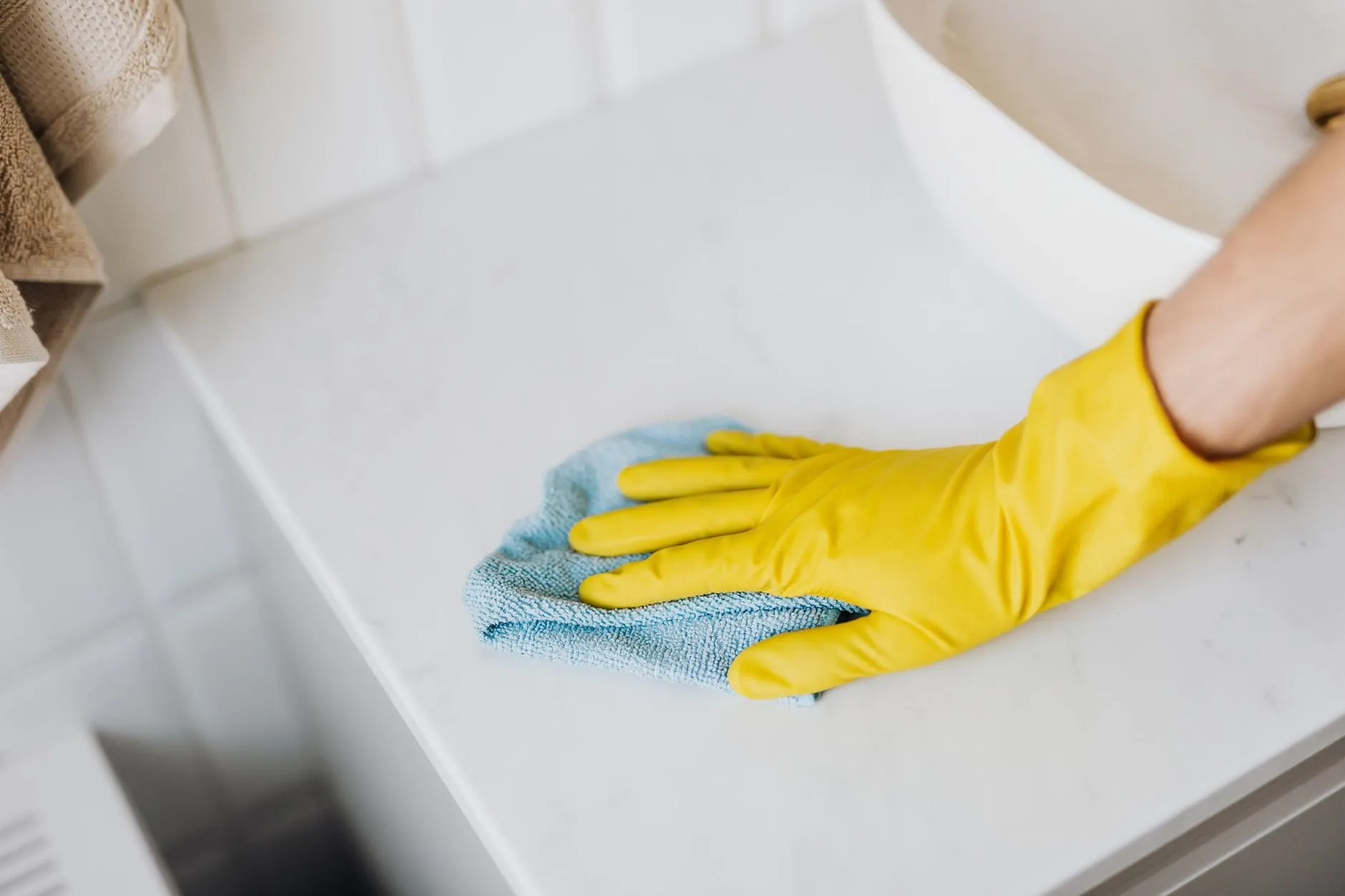 A gloved hand wiping a white kitchen worktop with a blue microfibre cloth